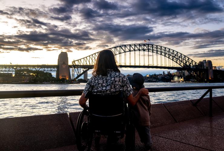 Ein Mann in einem Rollstuhl, während er mit seinem Sohn auf die Sydney Harbour Bridge blickt, Circular Quay, Sydney, New South Wales © Tourism Australia