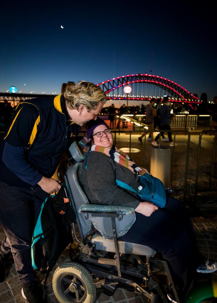 Eine Frau im Rollstuhl mit einer weiteren Person, die hinter ihr steht, und der Sydney Harbour Bridge im Hintergrund, Vivid, Sydney, New South Wales © Destination NSW
