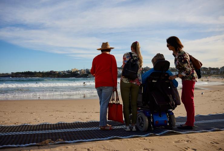 Ein Mann in einem Rollstuhl auf Strandmatten mit seiner Familie am Bondi Beach mit Australia In Style, Sydney, New South Wales © Destination NSW