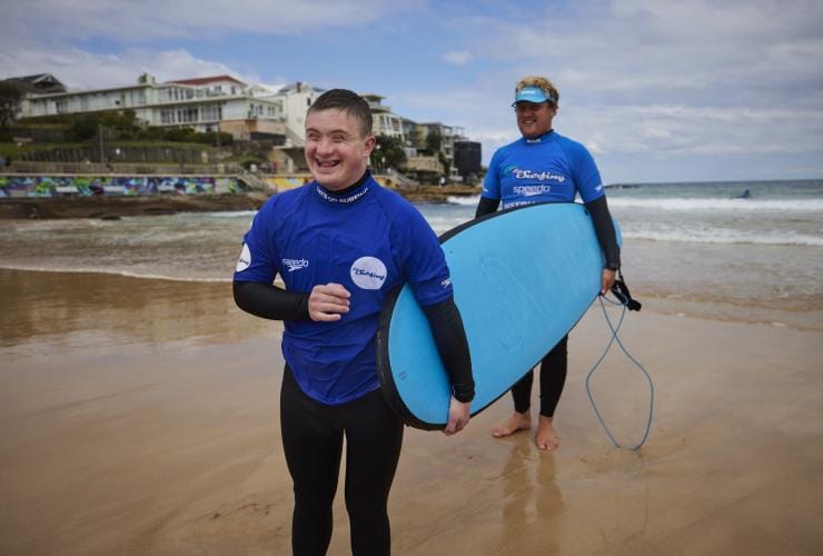 Seorang pria penyandang disabilitas saraf memegang papan selancar di pasir dengan instruktur dari Let's Go Surfing, Bondi Beach, Sydney, New South Wales © Tourism Australia