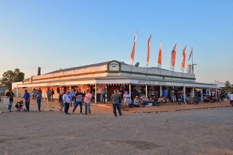 Ein historisches Pub mit umlaufender, mit Fahnen geschmückter Markise und Menschen, die draußen sitzen, trinken und sich unterhalten, Birdsville Hotel, Birdsville, Queensland © Tourism and Events Queensland