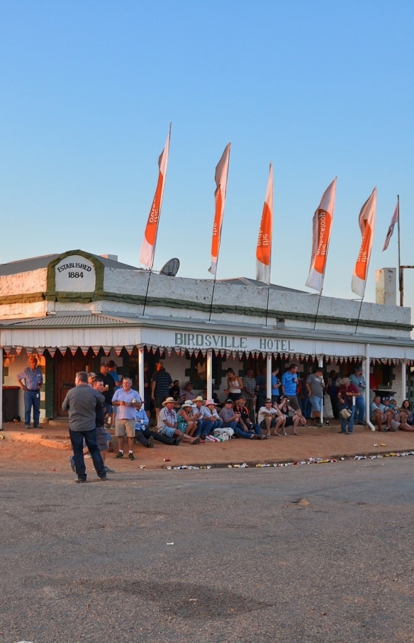 Ein historisches Pub mit umlaufender, mit Fahnen geschmückter Markise und Menschen, die draußen sitzen, trinken und sich unterhalten, Birdsville Hotel, Birdsville, Queensland © Tourism and Events Queensland