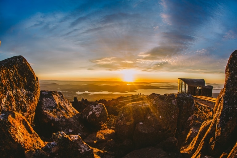 Große Felsen im Vordergrund mit zwei Personen, die an einem Aussichtspunkt in der Ferne stehen und den Blick über Hobart bewundern, während die Sonne über die Landschaft strahlt, kunanyi/Mt Wellington, Hobart, Tasmanien © Tourism Australia