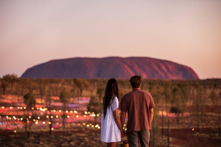 Ein Paar steht im Northern Territory bei Sonnenuntergang vor Uluru und dem Field of Light © Tourism NT/The Salty Travellers