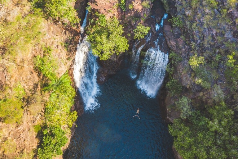 Luftaufnahme einer Person, die sich in einem Naturpool mit blauem Wasser treiben lässt, der von Bäumen umgeben ist und von zwei großen Wasserfällen gespeist wird, Florence Falls, Litchfield National Park, Northern Territory © Tourism NT/Dan Moore