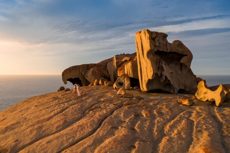 Zwei Personen stehen bei Sonnenuntergang mit dem Ozean im Hintergrund neben den riesigen Formationen der Remarkable Rocks auf Kangaroo Island, Südaustralien © South Australian Tourism Commission