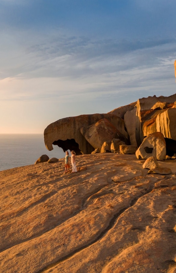 Zwei Personen stehen bei Sonnenuntergang mit dem Ozean im Hintergrund neben den riesigen Formationen der Remarkable Rocks auf Kangaroo Island, Südaustralien © South Australian Tourism Commission