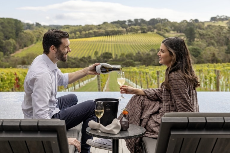 Ein Mann schenkt einer Frau ein Glas Weißwein ein, während sie auf Stühlen sitzen, mit einem Weinberg im Hintergrund, Jackalope Hotel, Mornington Peninsula, Victoria © Tourism Australia