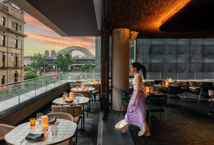 A person walking across the balcony of a restaurant with candle-lit tables overlooking a view of a large, steel arch bridge during a colourful pink sunset. Penelope's, Sydney, New South Wales © Penelope's/Chad Konik