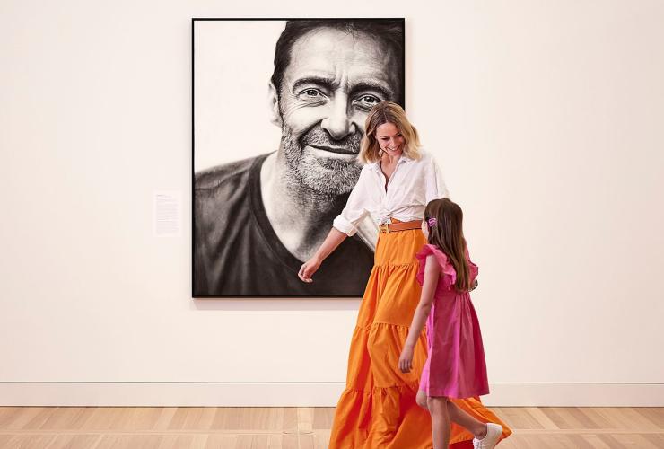 A mother and daughter wearing bright pink and orange clothes walking past a black and white painting of a man smiling against a blank wall at the National Portrait Gallery, Canberra, Australian Capital Territory © Crux for VisitCanberra