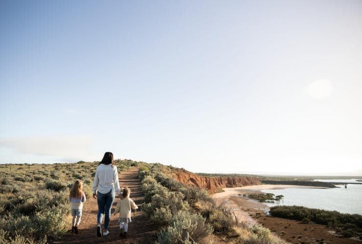 A woman and two children walking on a path bordered by bushland along a headland overlooking a calm blue waterway at Australian Arid Lands Botanic Garden, Port Augusta, South Australia © Tourism Australia / South Australian Tourism Commission    