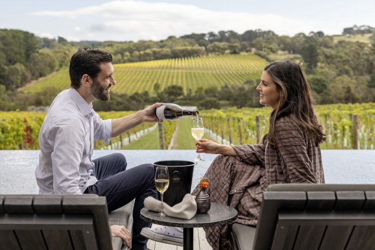 A man pouring a woman a glass of white wine as they sit on chairs facing a vineyard at Jackalope Hotel, Mornington Peninsula, Victoria © Tourism Australia