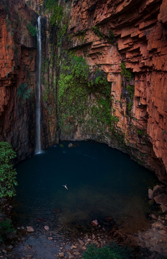 Aerial view over a person floating on their back in a blue waterhole surrounded by towering red rock walls adorned with moss and a slim waterfall at Emma Gorge, El Questro Wilderness Park, Kimberley, Western Australia © Tourism Australia