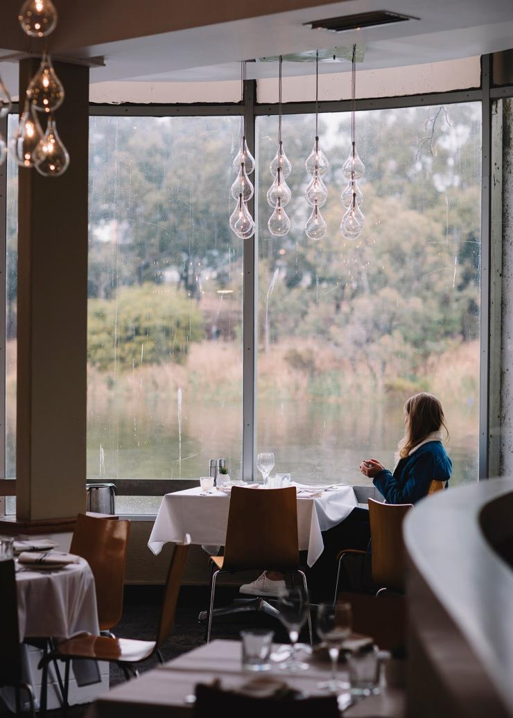A person sitting in a restaurant beside a floor to ceiling window admiring the view over a tranquil waterway at the River Café, North Adelaide, South Australia © Tourism Australia