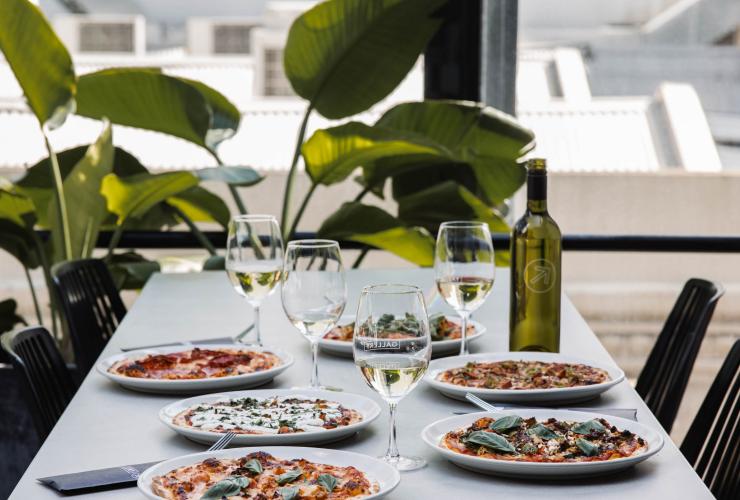 A table beside a window and green, leafy plant set with an assortment of pizzas and glasses of white wine at  Gallery Bar, Adelaide, South Australia © Lewis Potter/Gallery Bar