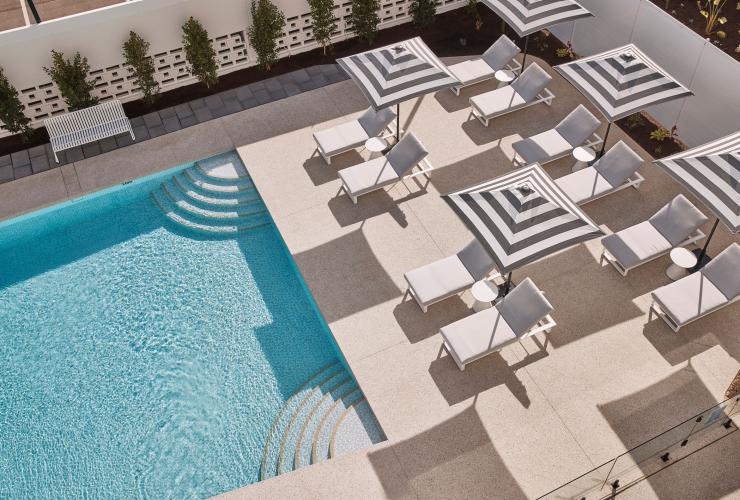 Aerial view over a blue pool surrounded by white tiles with white lounge chairs shaded by striped umbrellas at Hotel Alba, Adelaide, South Australia © South Australian Tourism Commission