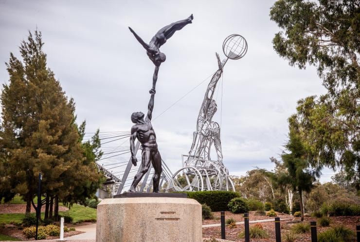 A statue in the foreground with two gymnasts holding hands and a larger statue in the background of a person in a wheelchair reaching for a basketball on a property surrounded by trees at Australian Institute of Sport, Canberra, Australian Capital Territory © Richard Poulton/VisitCanberra