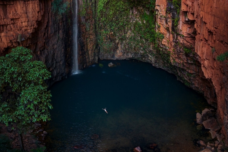 Aerial view over a person floating on their back in a blue waterhole surrounded by towering red rock walls adorned with moss and a slim waterfall at Emma Gorge, El Questro Wilderness Park, Kimberley, Western Australia © Tourism Australia