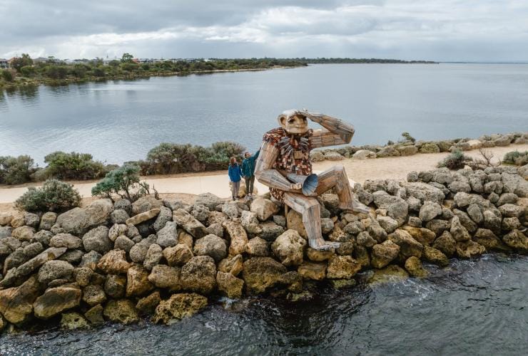 Aerial view over a rock-lined pier over a tranquil waterway, with two people standing beside a giant sculpture that appears to be sitting on the edge and looking out to sea at Seba's Song Giant, Giants of Mandurah by Thomas Dambo, Mandurah, Western Australia © Visit Mandurah
