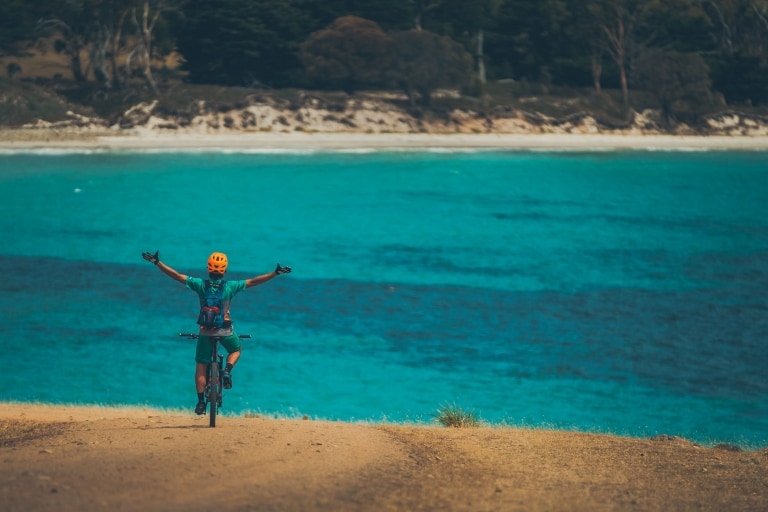 A person on a mountain bike with their arms in the air overlooking clear blue ocean in Maria Island National Park, Tasmania © Matt Staggs