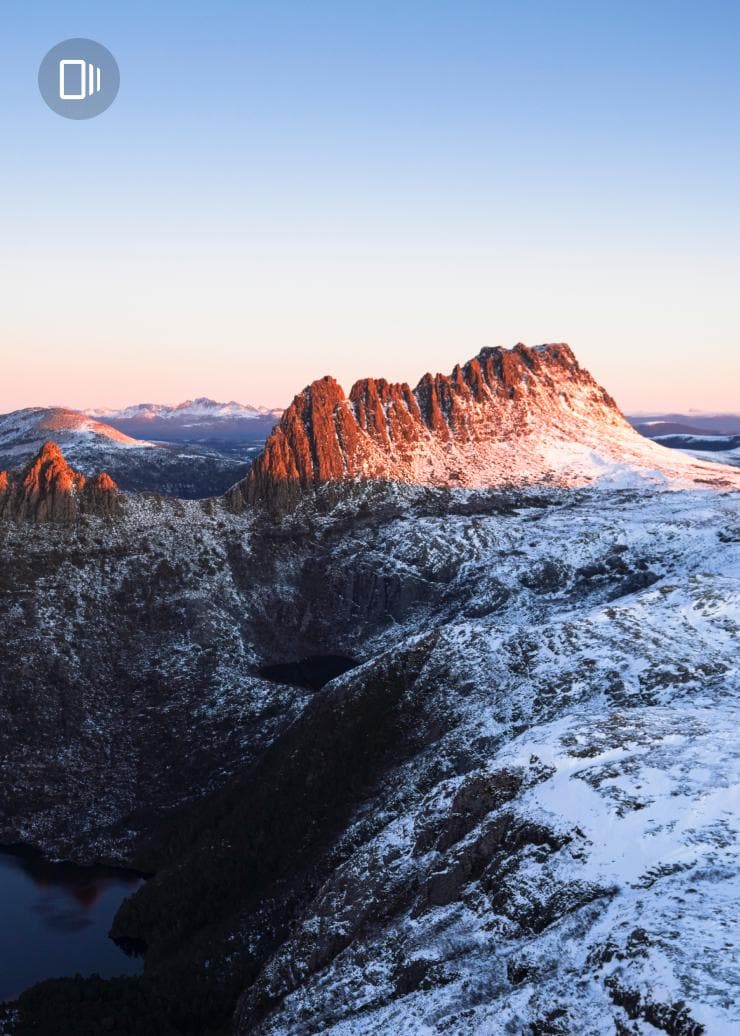 A rocky mountain range dusted with snow beneath a clear blue sky, with peaks glowing orange in the sunlight, Tasmania © Luke Tscharke