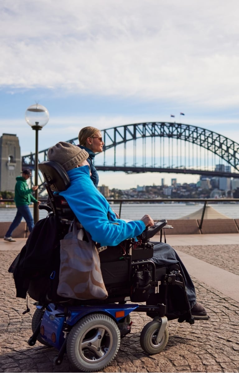 Person using a mobility device with another person walking beside them approaching the Sydney Opera House with the Sydney Harbour Bridge in the background in Sydney, New South Wales © Destination NSW