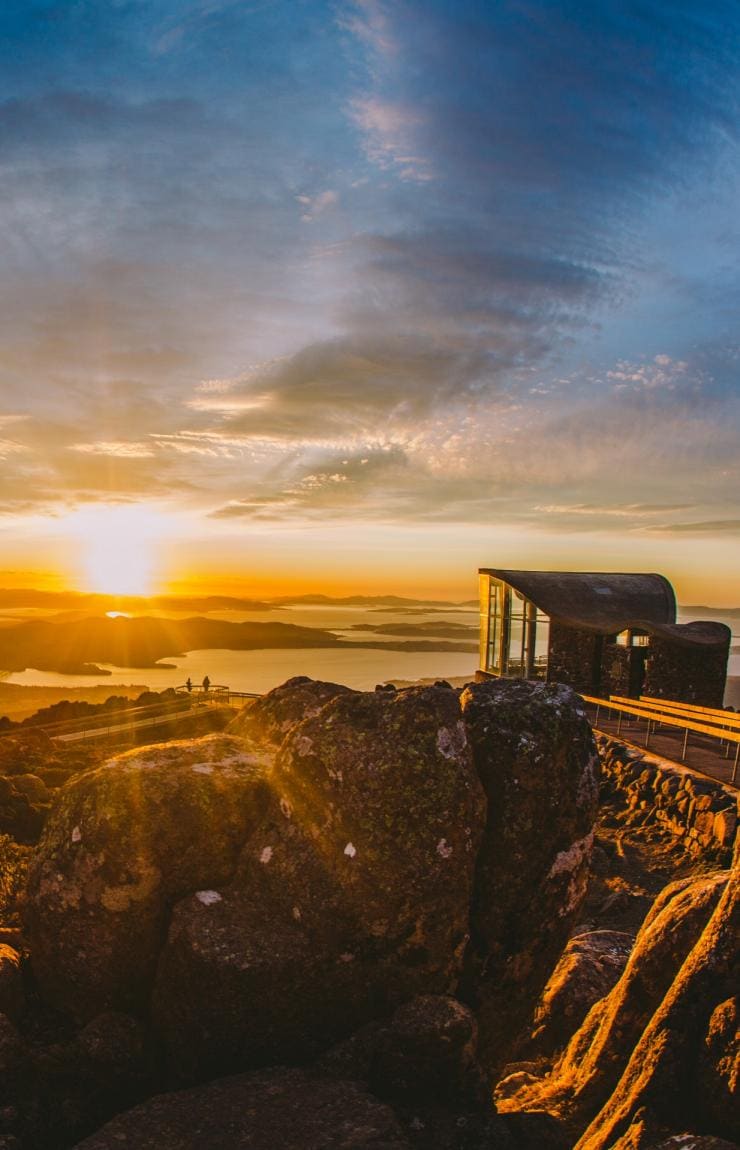 Large rocks in the foreground with two people standing at a lookout in the distance admiring views over Hobart as the sun beams over the landscape at kunanyi/Mt Wellington, Hobart, Tasmania © Tourism Australia