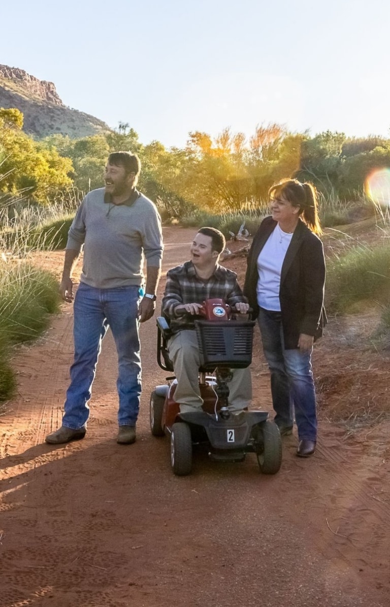 Seorang Pria duduk di skuter elektronik di samping pria lain dan seorang wanita tengah berjalan di antara tanaman hijau dan tanah merah di Alice Springs Desert Park di Alice Springs, Northern Territory