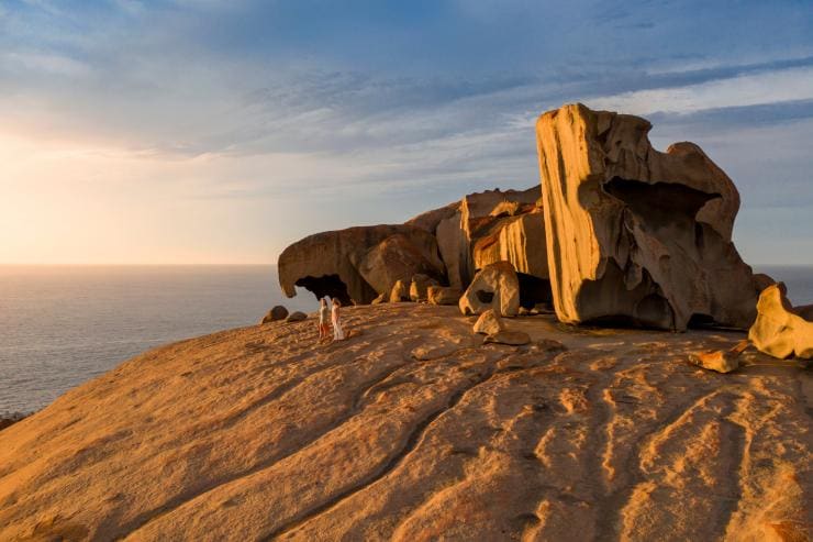 Dua orang sedang berjalan di samping formasi besar Remarkable Rocks saat matahari terbenam dengan latar belakang samudra di Kangaroo Island, South Australia © South Australian Tourism Commission