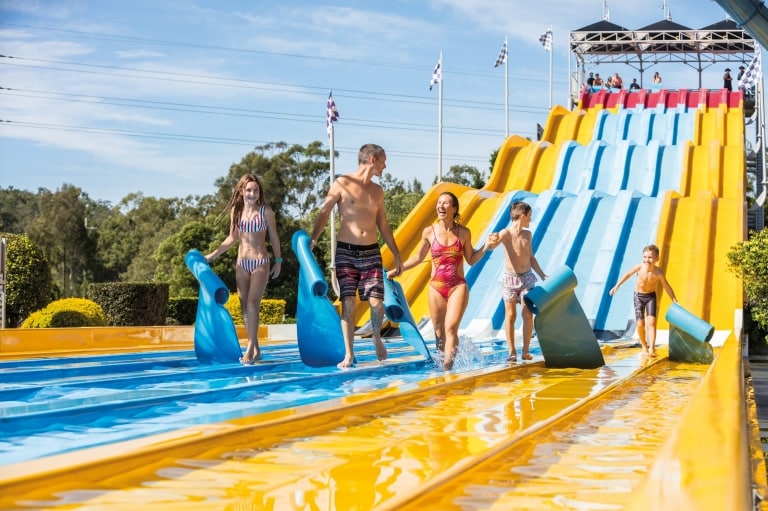 クイーンズランド州ゴールド・コースト、ウェット・アンド・ワイルド・ウォーターパークにて、黄・青・赤のスライダーを滑り終えて笑顔で歩く家族 © Destination Gold Coast