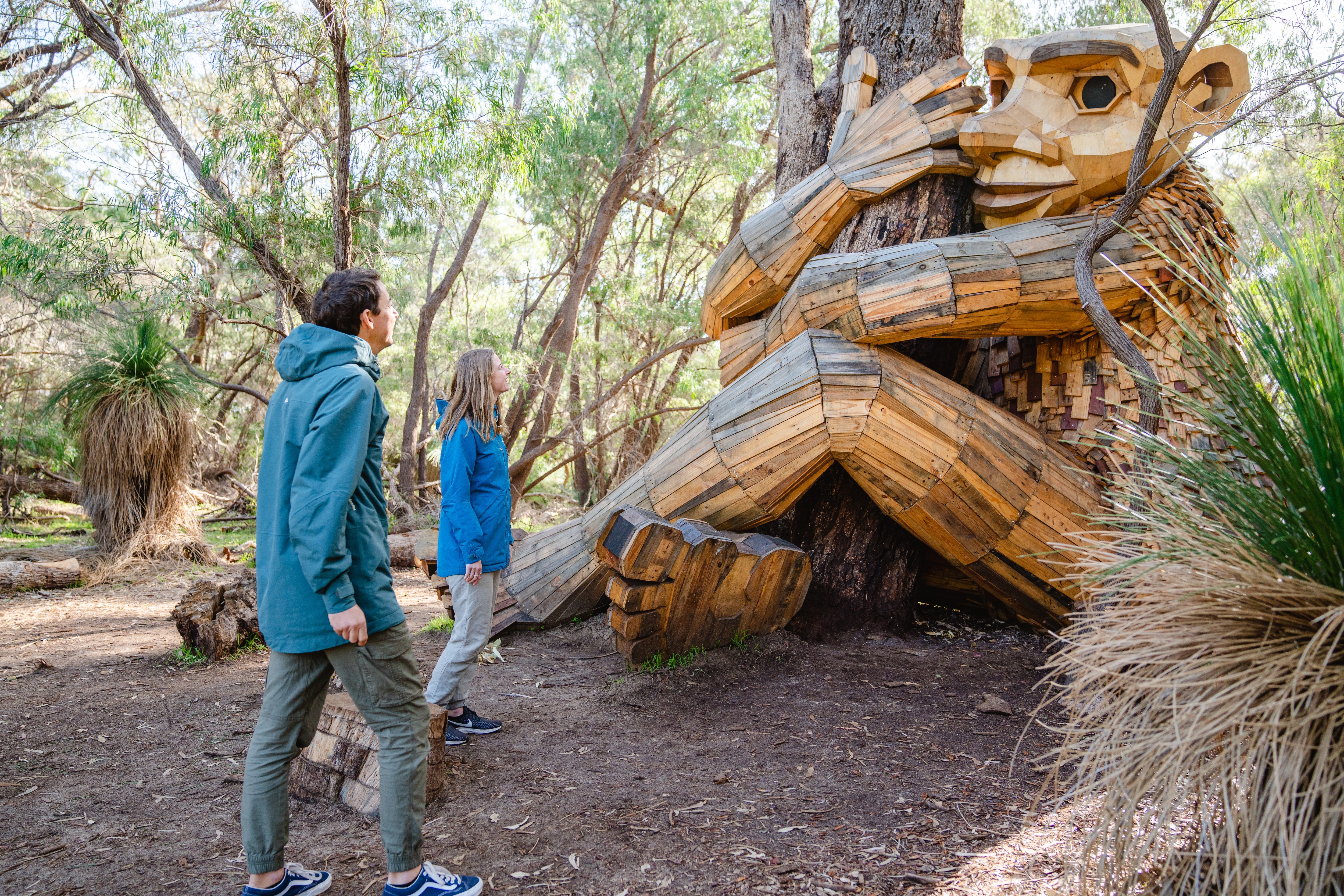 Two people approaching a large wooden sculpture of a giant hugging a tree at Giants of Mandurah by Thomas Dambo, Mandurah, Western Australia © Visit Mandurah