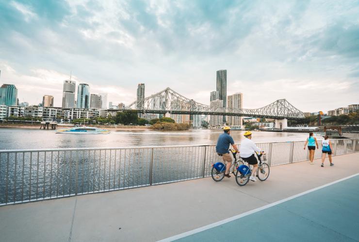 Spaziergänger und Radfahrer auf einem Weg entlang des Brisbane River mit der Skyline der Stadt in der Ferne, River Walk, Brisbane, Queensland © Tourism and Events Queensland