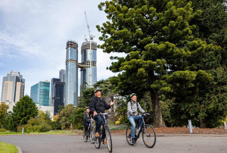 Drei Radfahrer fahren während einer Tour mit Melbourne by Bike auf einem ruhigen Weg durch einen grünen Park mit der Skyline der Stadt im Hintergrund, Melbourne, Victoria © Tourism Australia