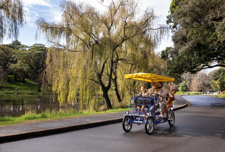 Eine Familie fährt auf einem vierrädrigen Fahrrad mit Sonnenschirm an einem Teich vorbei durch die Grünanlagen von Centennial Park Cycles, Centennial Park, New South Wales © Destination NSW