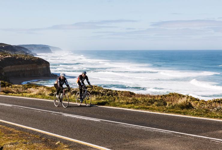 Zwei Radfahrer fahren nebeneinander auf einer Straße an steilen Sandsteinklippen entlang, die zum Meer hin abfallen, Great Ocean Road, Victoria © Belinda Van Zanen/Great Ocean Road Tourism