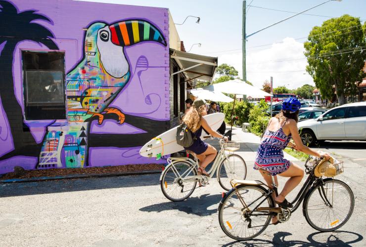 Zwei Radfahrerinnen, von denen eine ein Surfbrett hält, fahren an einem farbenfrohen Wandgemälde eines Tukans vorbei, Byron Bay, New South Wales © Destination NSW/James Horan