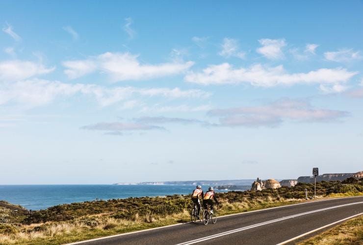 Zwei Radfahrer fahren nebeneinander an einem Klippenrand entlang auf einer Straße, die zum Ozean führt, mit den 12 Apostles in der Ferne, Great Ocean Road, Victoria © Belinda Van Zanen/Great Ocean Road Tourism