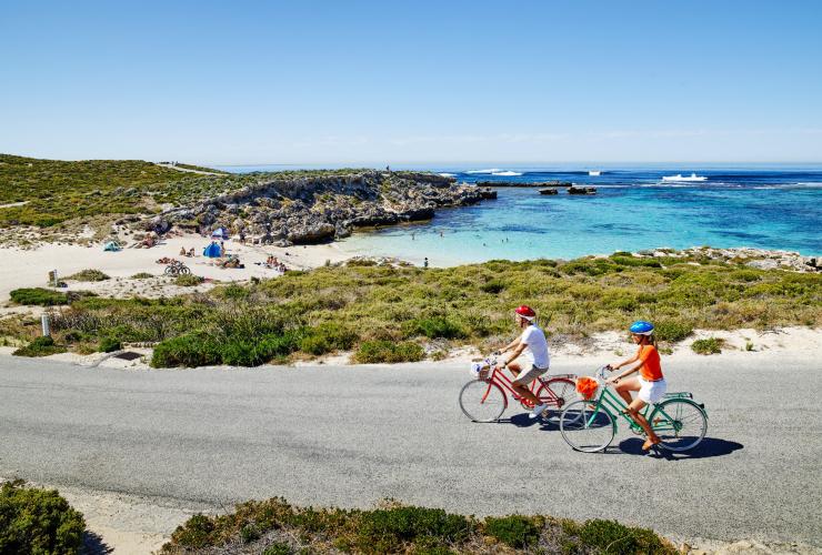 Zwei Radfahrer fahren auf bunten Fahrrädern auf einer Straße entlang einer Bucht mit weißem Sand und klarem blauen Wasser, Little Salmon Bay, Rottnest Island, Westaustralien © Tourism Western Australia
