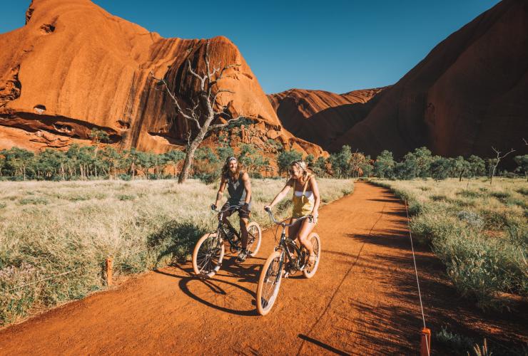 Zwei Radfahrer fahren lachend auf einem roten Sandweg mit der hoch aufragenden Formation des Uluru hinter sich, Uluru-Kata Tjuta National Park, Northern Territory © Tourism NT/Laura Bell