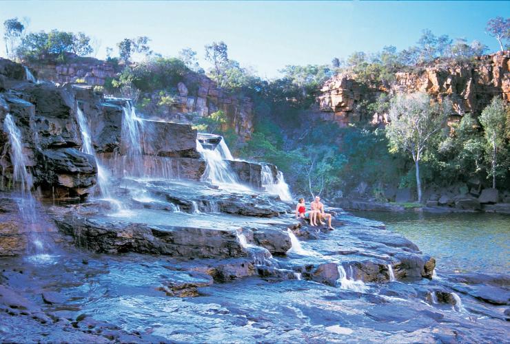 Zwei Personen sitzen auf einem Felsen mit Stufen und Wasserfall an der Gibb River Road, Manning Gorge Waterfall, Mount Barnett Station, Westaustralien © Tourism Western Australia