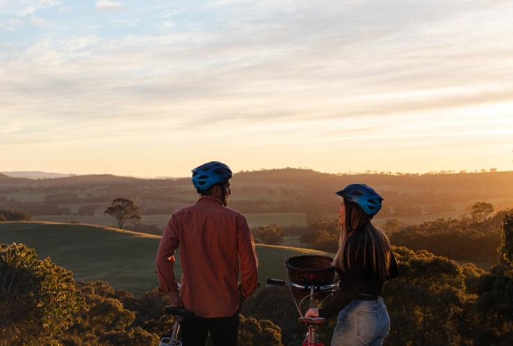 Ein Mann und eine Frau stehen neben Fahrrädern und blicken bei Sonnenuntergang auf sanfte Hügel, Paulett Wines, Clare Valley, Südaustralien © South Australian Tourism Commission/Harry Vick
