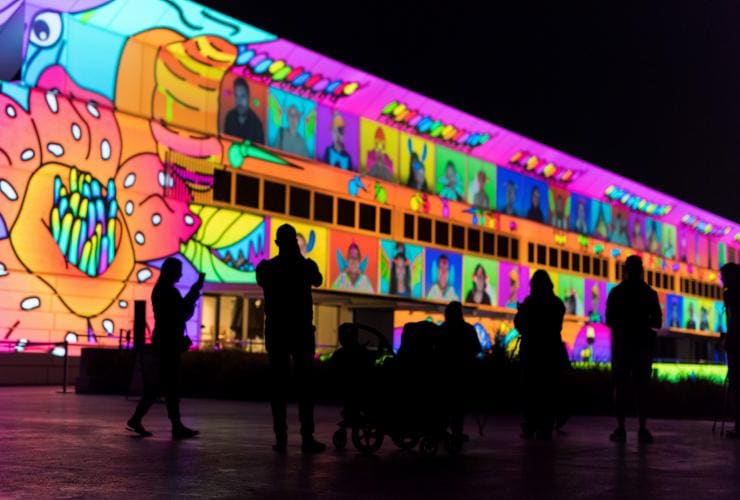 Eine Gruppe von Menschen wirft dunkle Silhouetten vor ein Gebäude, das in einer farbenfrohen Kunstinstallation namens Illuminations beleuchtet wird, Enlighten Festival, Canberra, Australian Capital Territory © Enlighten Festival