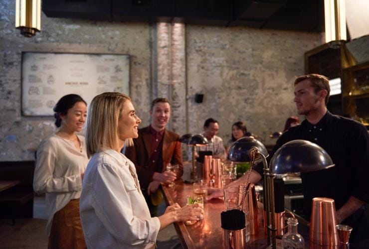 Eine Gruppe lächelnder Personen steht mit Getränken an der Bar in der Archie Rose Distillery, Sydney, New South Wales © Destination NSW