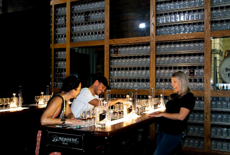Drei Personen stehen lachend um einen Tisch mit Gläsern, die mit Rum gefüllt sind, Bundaberg Rum Experience, Bundaberg, Queensland © Tourism and Events Queensland