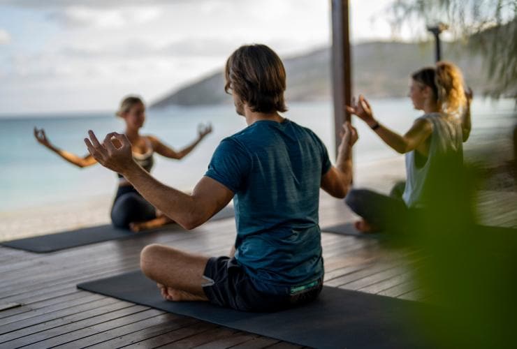 Ein Paar sitzt unter einer Cabana am Strand und nimmt unter Anleitung einer Yogalehrerin eine Yogastellung ein, Lizard Island Resort, Lizard Island, Queensland © Tourism and Events Queensland