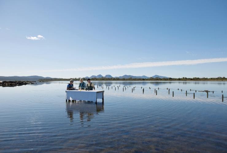 Ein Paar speist an einem Tisch im Wasser auf der Freycinet Marine Oyster Farm während einer Saffire Signature Experience, Coles Bay, Freycinet, Tasmanien © Tourism Tasmania/George Apostolidis