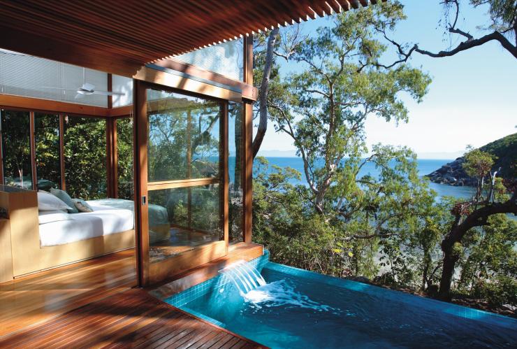 Eine Holzvilla mit einem Bett in einem von Fenstern umgebenen Raum neben einem Infinity-Pool mit Blick auf das Buschland und den Ozean darunter, Bedarra Island, Queensland © Bedarra Island