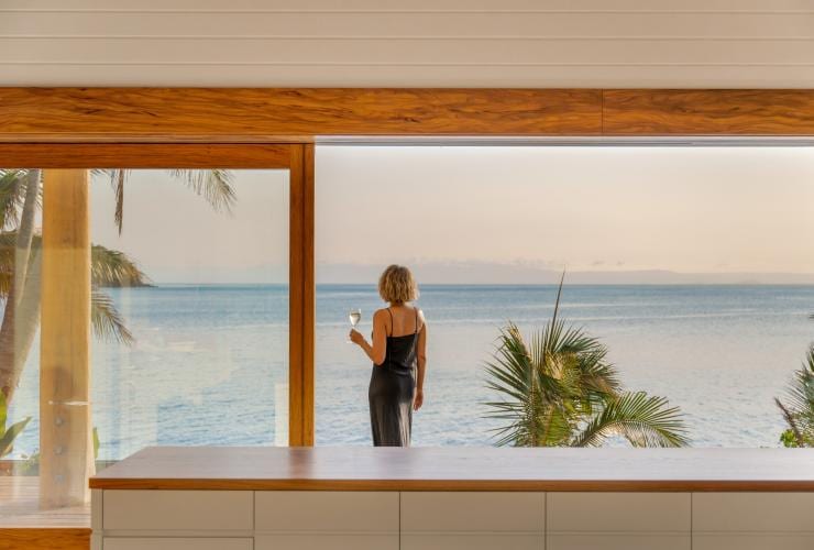 Eine Person steht mit einem Glas Weißwein in der Hand auf einem Balkon und blickt auf den Ozean, Pelorus Private Island, Palm Island, Queensland © James Vodicka/Pelorus Private Island