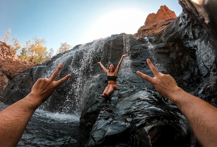 A woman sitting in a waterfall as someone near the camera gives a peace sign at Edith Falls, Nitmiluk National Park, Northern Territory © Tourism Australia