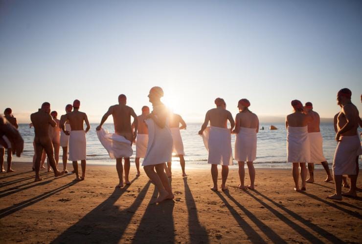 A group of people stand on the beach wearing towels at the Nude Solstice Swim, Dark Mofo, Hobart, Tasmania © Dark Mofo